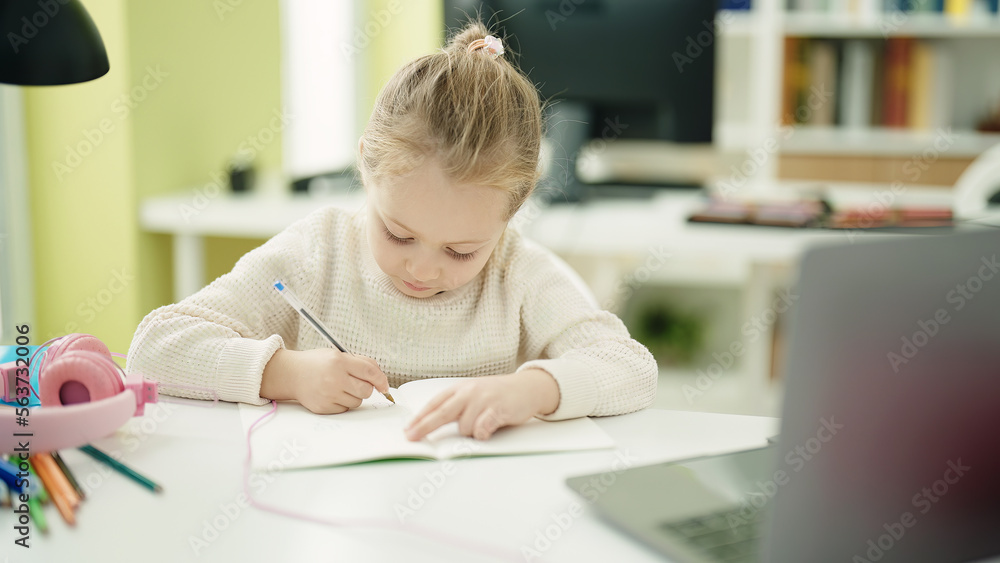 Adorable blonde girl student writing on notebook sitting on table at classroom Adorable blonde girl student writing on notebook sitting on table at classroom