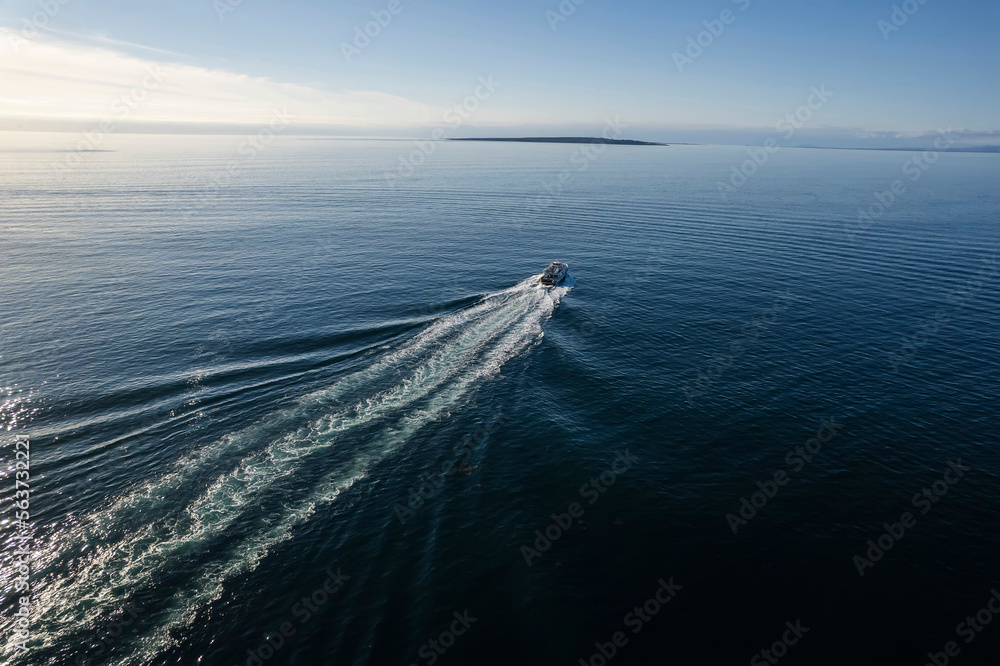 Small ferry boat in blue ocean on the way to Aran island, Ireland. Wake behind cruise ship. Warm sunny day with clear blue sky. Travel and transportation industry. Popular route from Doolin port.