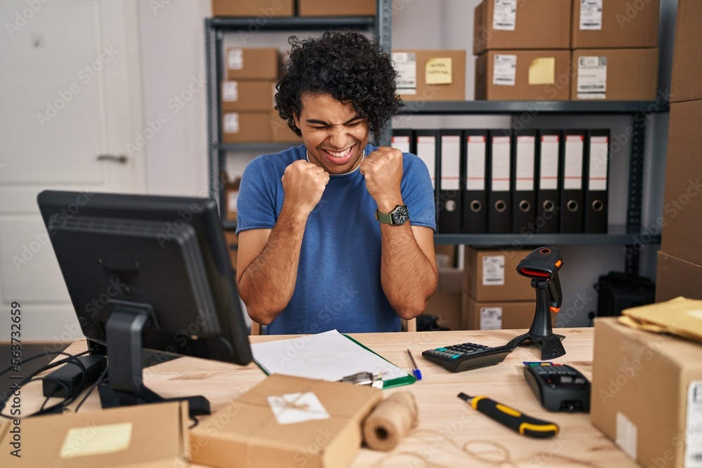 Hispanic man with curly hair working at small business ecommerce excited for success with arms raised and eyes closed celebrating victory smiling. winner concept. Hispanic man with curly hair working at small business ecommerce excited for success with arms raised and eyes closed celebrating victory smiling. winner concept.