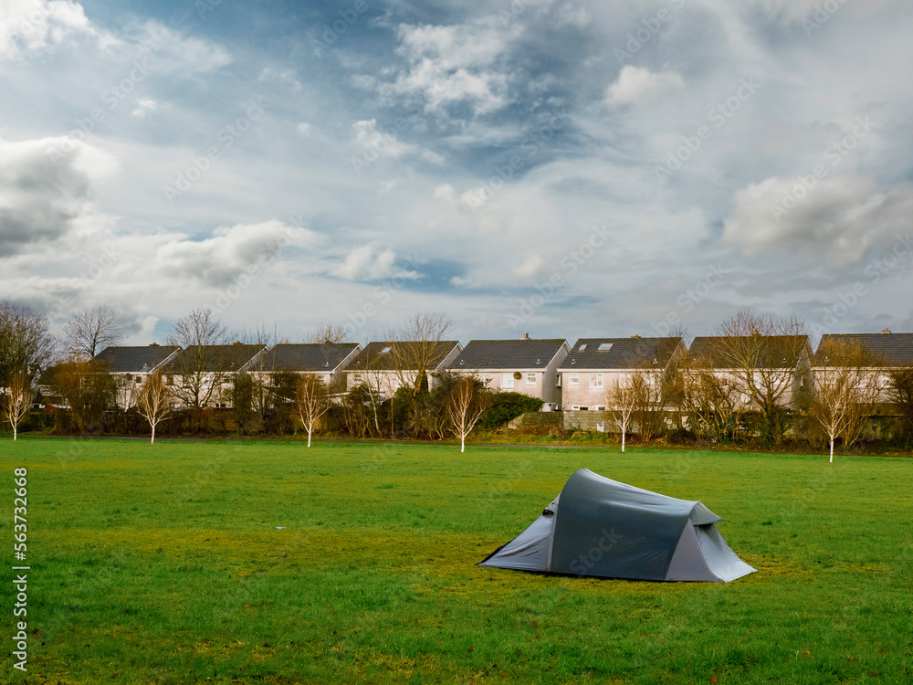 Simple tent on green grass in a park. Row of houses in the background. Stunning blue cloudy sky. Homeless living close to dense populated area. Social issue.