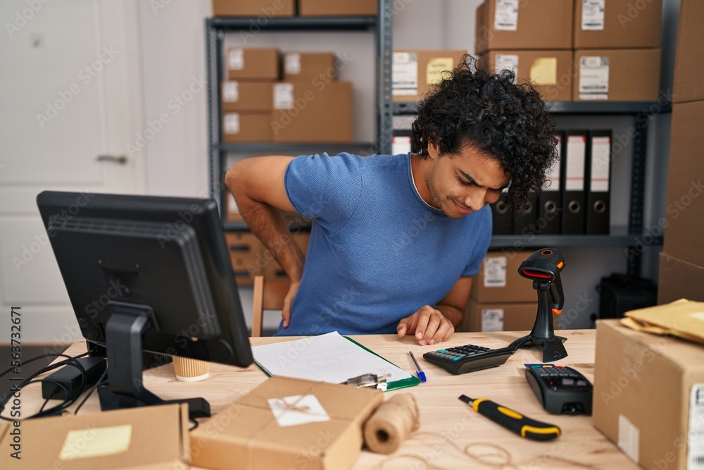 Hispanic man with curly hair working at small business ecommerce suffering of backache, touching back with hand, muscular pain Hispanic man with curly hair working at small business ecommerce suffering of backache, touching back with hand, muscular pain