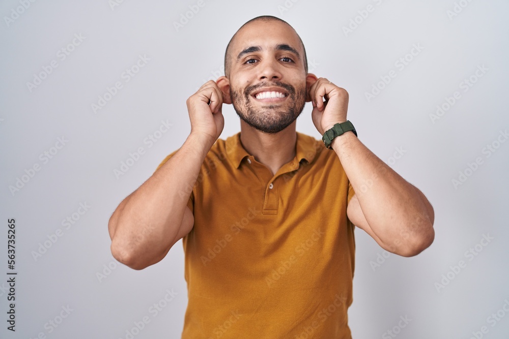 Fototapeta premium Hispanic man with beard standing over white background smiling pulling ears with fingers, funny gesture. audition problem