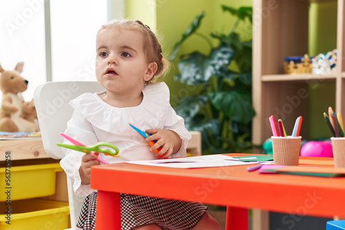 Adorable blonde toddler student sitting on table holding scissors at kindergarten