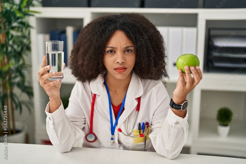 Young african american dietitian woman holding fresh apple and water clueless and confused expression. doubt concept. Young african american dietitian woman holding fresh apple and water clueless and confused expression. doubt concept.