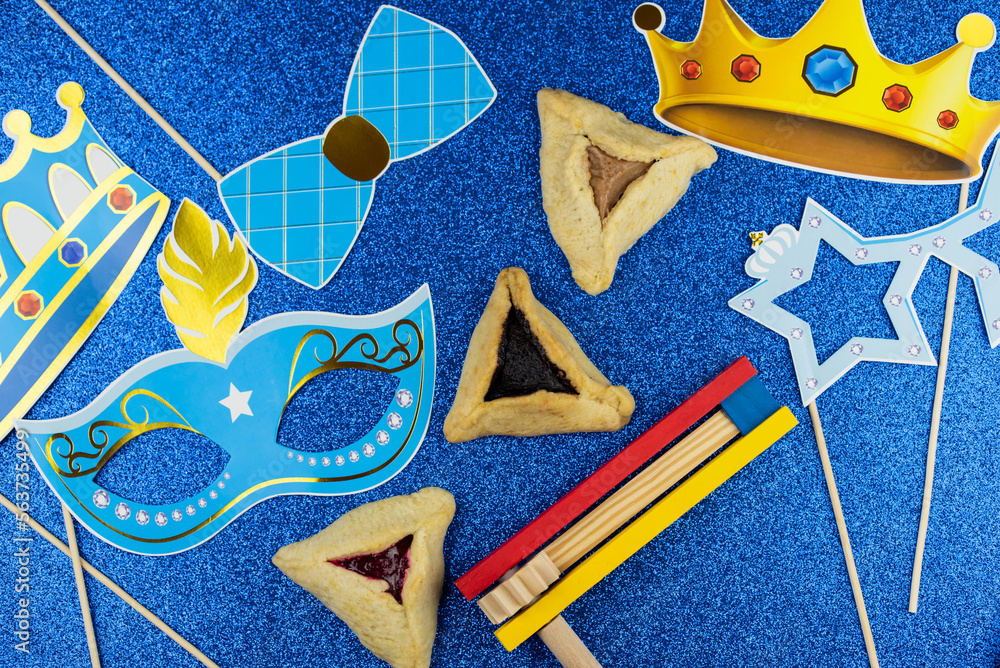 Jewish holiday Purim celebration concept. Traditional hamantasch or hamantaschen Haman's ears cookies, carnival mask, spin gragger (ratchet) noisemaker, and party decor on blue background.