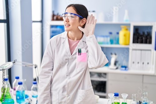Young brunette woman working at scientist laboratory smiling with hand over ear listening and hearing to rumor or gossip. deafness concept.