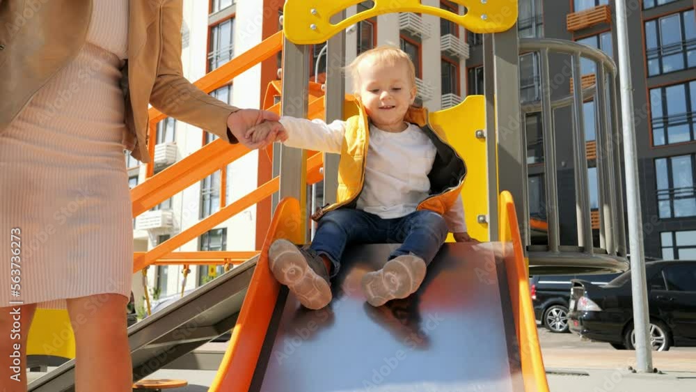 Slow motion of little baby boy with mother riding down the slide on playground. Happy parenting, family having time together, kids and parents outdoors
