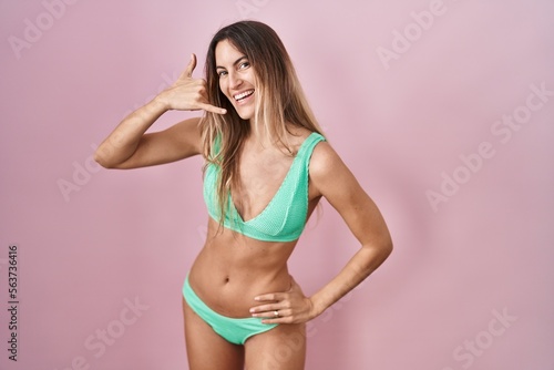 Young hispanic woman wearing bikini over pink background smiling doing phone gesture with hand and fingers like talking on the telephone. communicating concepts.