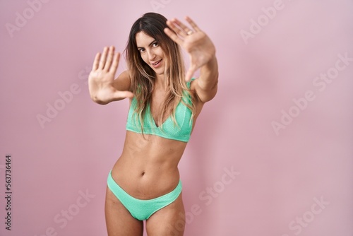 Young hispanic woman wearing bikini over pink background doing frame using hands palms and fingers, camera perspective © Krakenimages.com