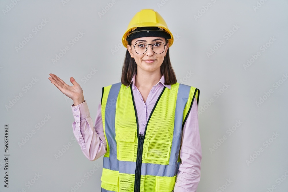 Hispanic girl wearing builder uniform and hardhat smiling cheerful presenting and pointing with palm of hand looking at the camera.