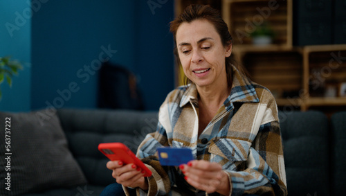 Middle age hispanic woman using smartphone and credit card sitting on sofa at home