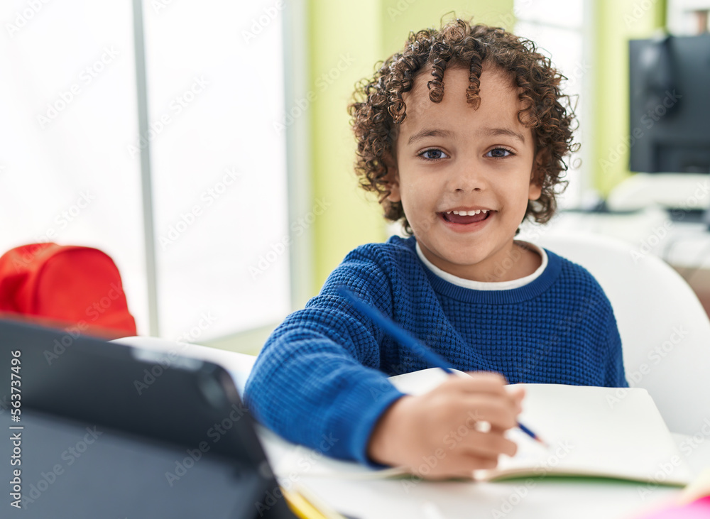 Adorable hispanic boy preschool student sitting on table drawing on notebook at classroom Adorable hispanic boy preschool student sitting on table drawing on notebook at classroom