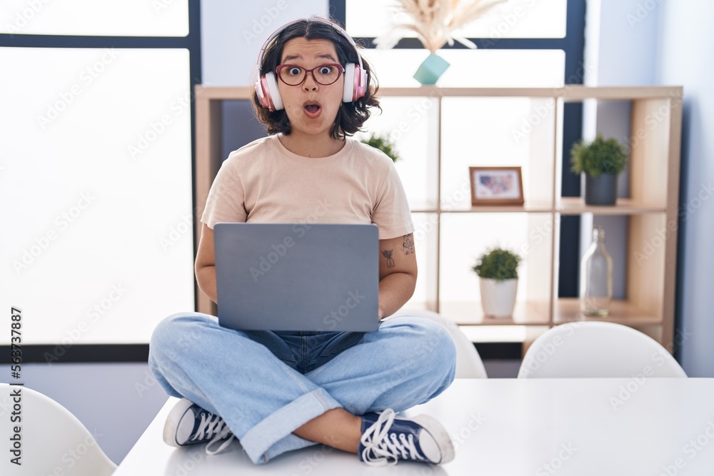 Young hispanic woman using laptop sitting on the table wearing headphones afraid and shocked with surprise expression, fear and excited face. Young hispanic woman using laptop sitting on the table wearing headphones afraid and shocked with surprise expression, fear and excited face.