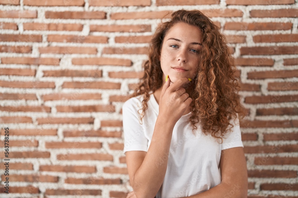 Young caucasian woman standing over bricks wall background looking confident at the camera with smile with crossed arms and hand raised on chin. thinking positive.