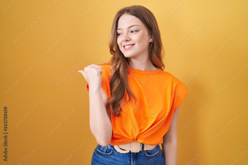 Caucasian woman standing over yellow background smiling with happy face looking and pointing to the side with thumb up. Caucasian woman standing over yellow background smiling with happy face looking and pointing to the side with thumb up.