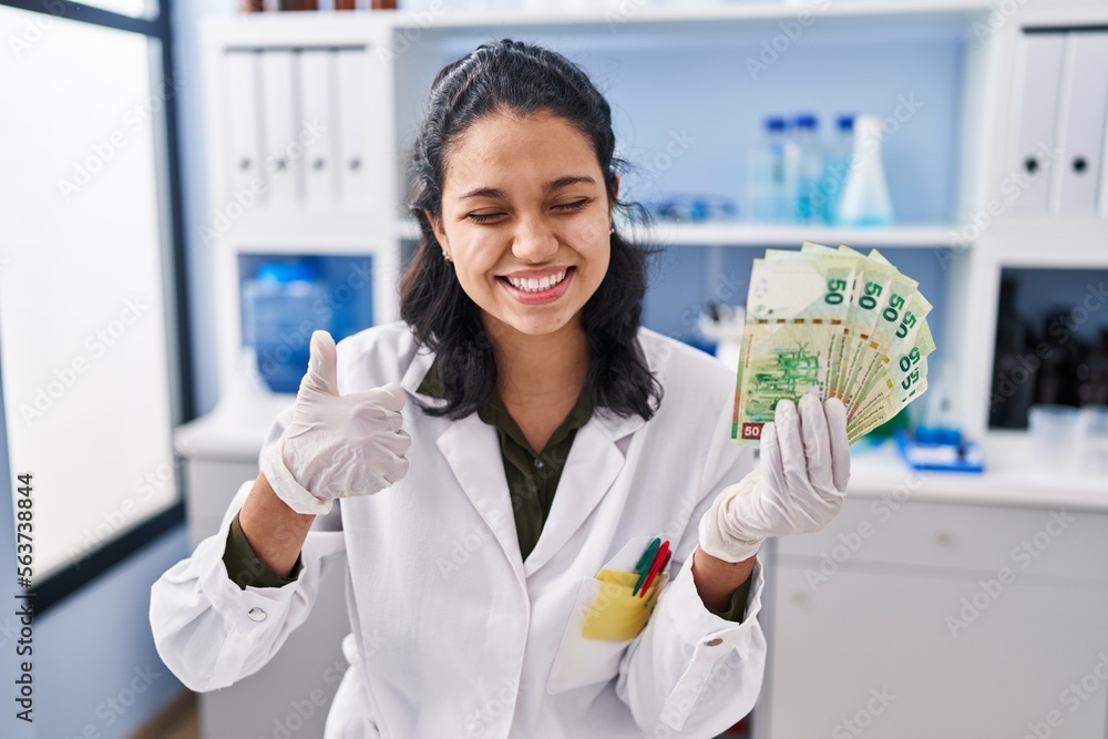 Hispanic woman with dark hair working at scientist laboratory holding money smiling happy and positive, thumb up doing excellent and approval sign Hispanic woman with dark hair working at scientist laboratory holding money smiling happy and positive, thumb up doing excellent and approval sign
