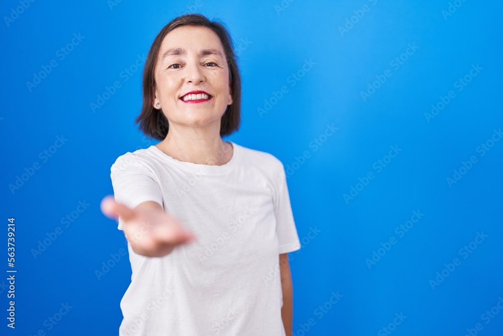 Middle age hispanic woman standing over blue background smiling cheerful offering palm hand giving assistance and acceptance. Middle age hispanic woman standing over blue background smiling cheerful offering palm hand giving assistance and acceptance.