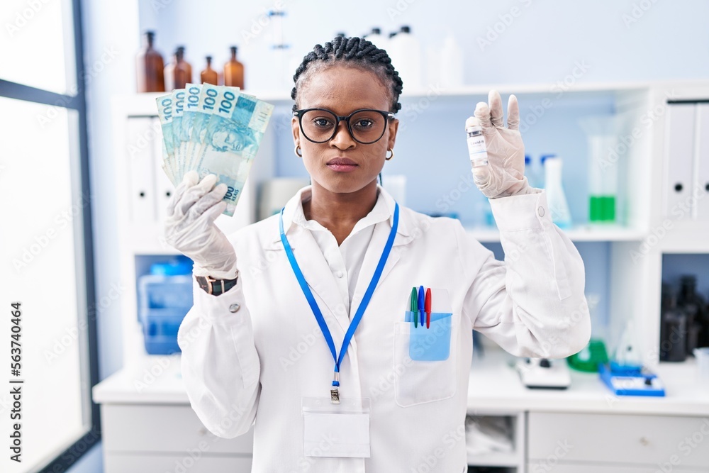 African woman with braids working at scientist laboratory holding money relaxed with serious expression on face. simple and natural looking at the camera. African woman with braids working at scientist laboratory holding money relaxed with serious expression on face. simple and natural looking at the camera.