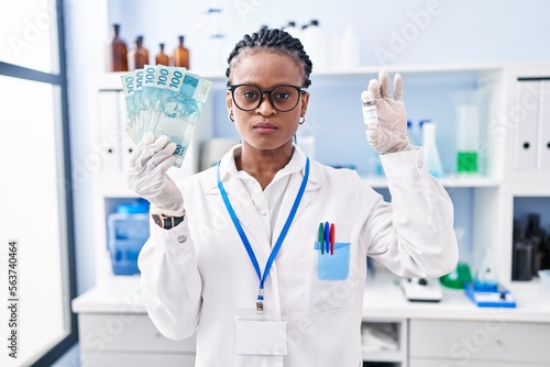 African woman with braids working at scientist laboratory holding money relaxed with serious expression on face. simple and natural looking at the camera.