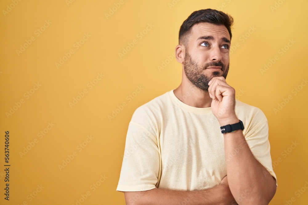 Handsome hispanic man standing over yellow background thinking worried about a question, concerned and nervous with hand on chin Handsome hispanic man standing over yellow background thinking worried about a question, concerned and nervous with hand on chin
