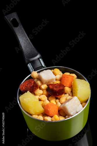 Cooked (Cocido, Guiso) stew, chickpeas with chorizo, bacon, carrot and potato in a green saucepan and on a black background. Concept of traditional Spanish cuisine.