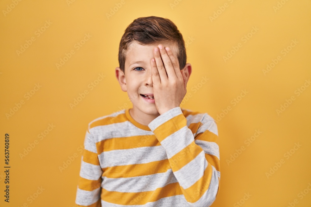 Young caucasian kid standing over yellow background covering one eye with hand, confident smile on face and surprise emotion. Young caucasian kid standing over yellow background covering one eye with hand, confident smile on face and surprise emotion.