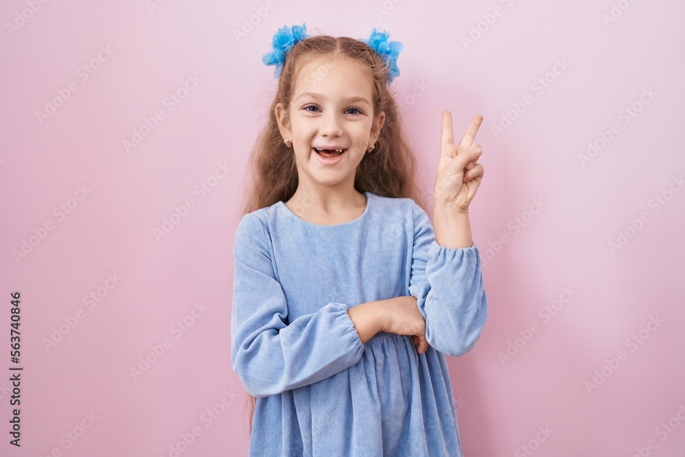Young little girl standing over pink background smiling with happy face winking at the camera doing victory sign. number two.