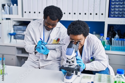 African american man and woman scientists using microscope writing on document at laboratory