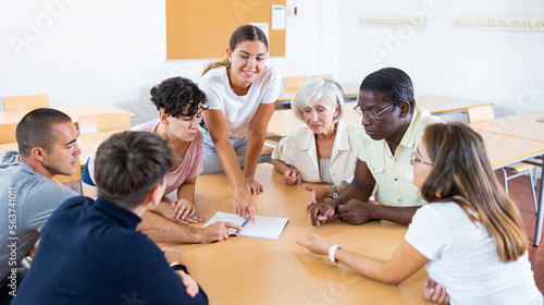 Photo Interested young girl participating in Spanish language speaking club, communicating with multiracial group of people of different ages sitting around table