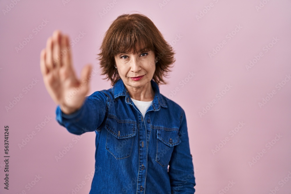 Middle age woman standing over pink background doing stop sing with palm of the hand. warning expression with negative and serious gesture on the face. Middle age woman standing over pink background doing stop sing with palm of the hand. warning expression with negative and serious gesture on the face.