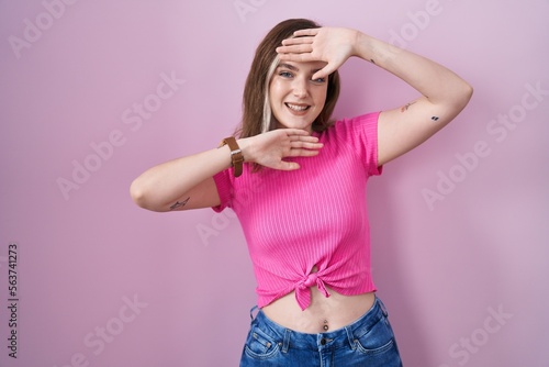 Blonde caucasian woman standing over pink background smiling cheerful playing peek a boo with hands showing face. surprised and exited