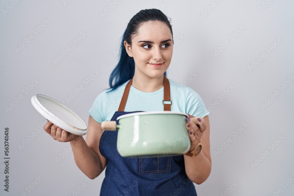 Young modern girl with blue hair wearing apron holding cooking pot smiling looking to the side and staring away thinking. Young modern girl with blue hair wearing apron holding cooking pot smiling looking to the side and staring away thinking.