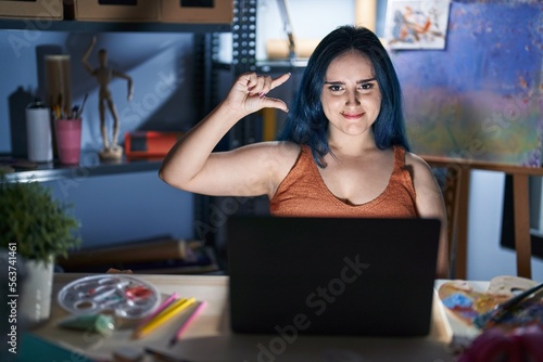 Young modern girl with blue hair sitting at art studio with laptop at night smiling and confident gesturing with hand doing small size sign with fingers looking and the camera. measure concept.