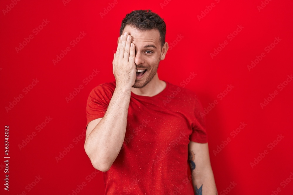 Young hispanic man standing over red background covering one eye with hand, confident smile on face and surprise emotion. Young hispanic man standing over red background covering one eye with hand, confident smile on face and surprise emotion.