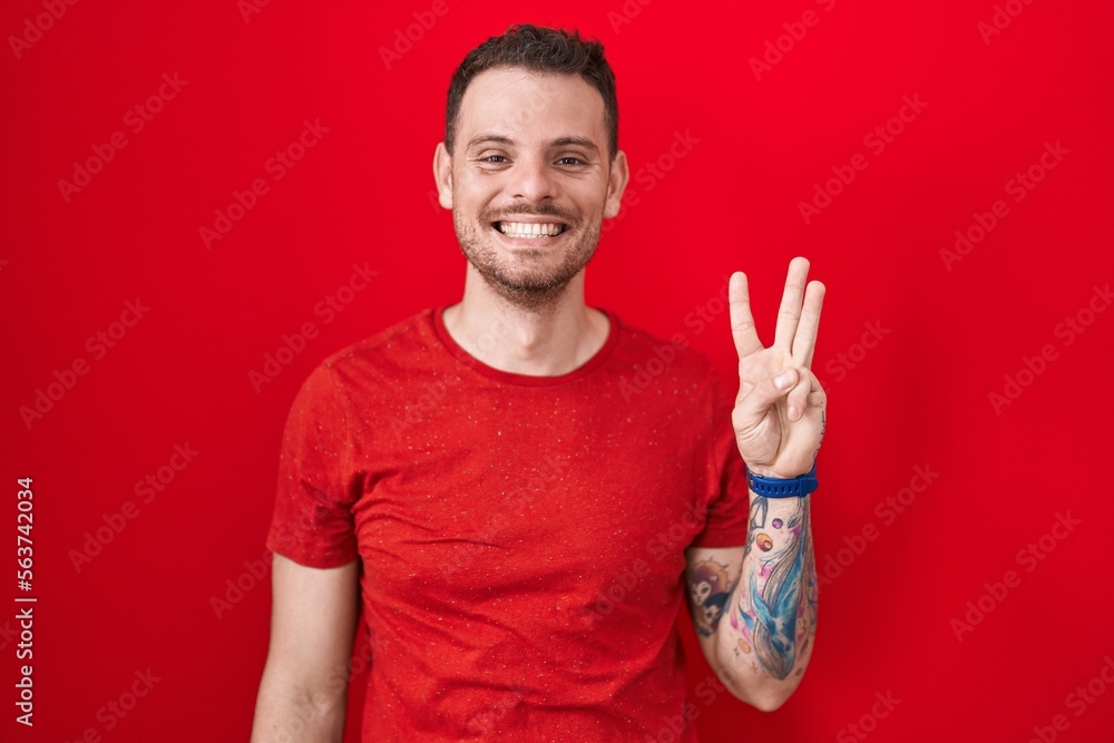 Young hispanic man standing over red background showing and pointing up with fingers number three while smiling confident and happy. Young hispanic man standing over red background showing and pointing up with fingers number three while smiling confident and happy.