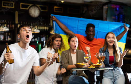 Happy international sport fans waving flag Ukraine during football match, drinking beer, eating chips in the pub