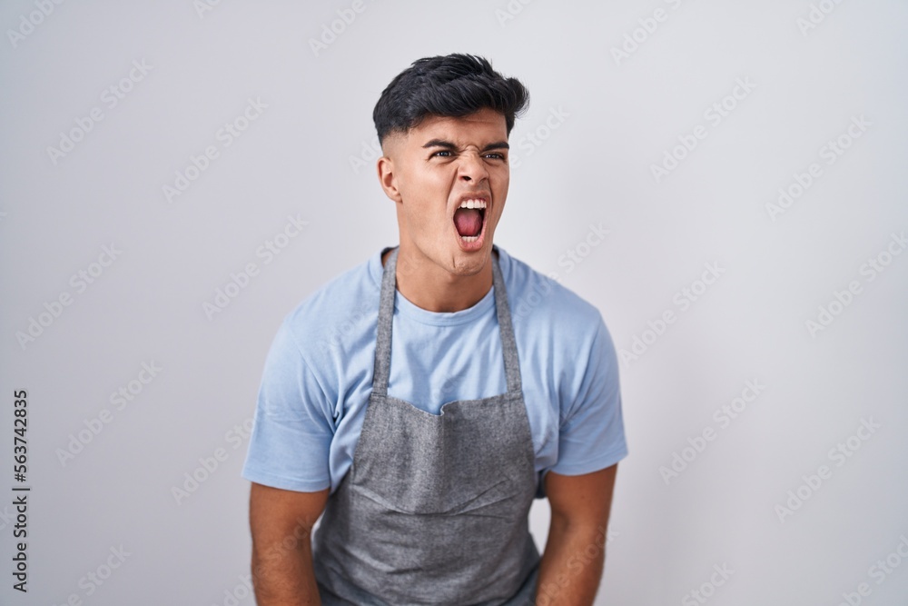 Hispanic young man wearing apron over white background angry and mad screaming frustrated and furious, shouting with anger. rage and aggressive concept. Hispanic young man wearing apron over white background angry and mad screaming frustrated and furious, shouting with anger. rage and aggressive concept.