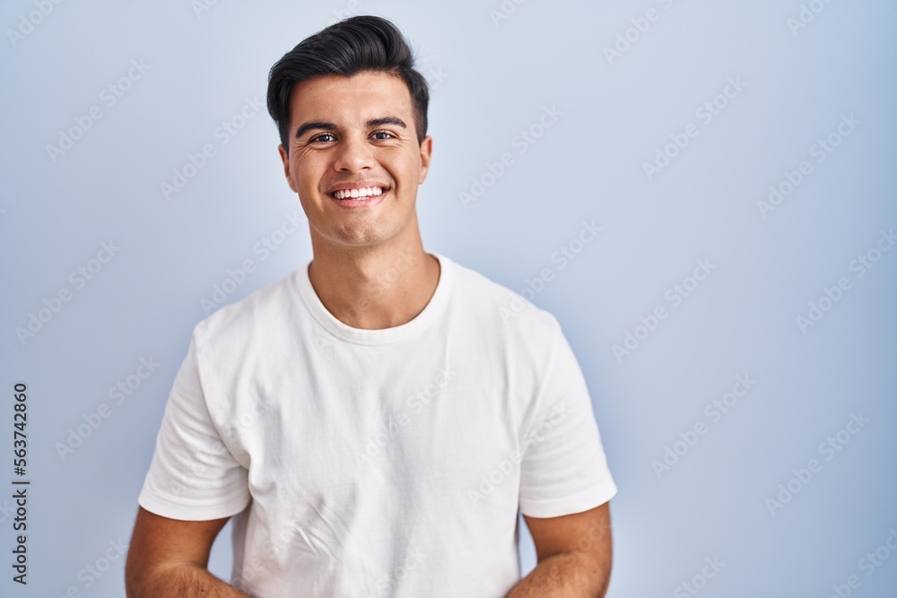 Hispanic man standing over blue background with hands together and crossed fingers smiling relaxed and cheerful. success and optimistic Hispanic man standing over blue background with hands together and crossed fingers smiling relaxed and cheerful. success and optimistic