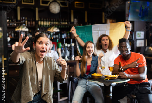 Excited young woman, Ireland football team fan, gesturing with hands, spending time in bar with friends. People with state flag in pub.