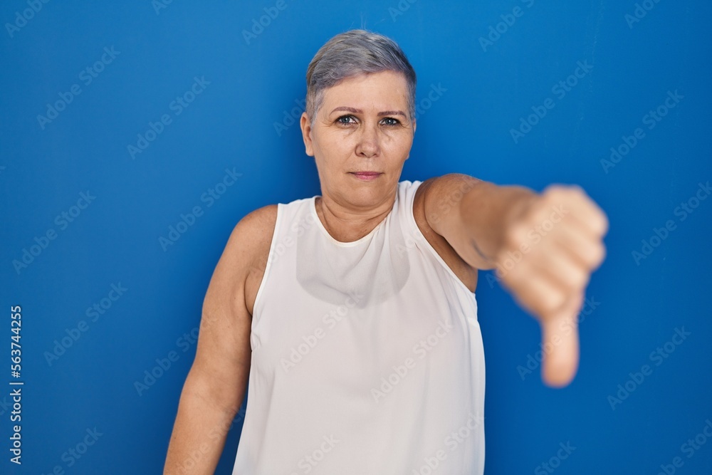 Fototapeta premium Middle age caucasian woman standing over blue background looking unhappy and angry showing rejection and negative with thumbs down gesture. bad expression.
