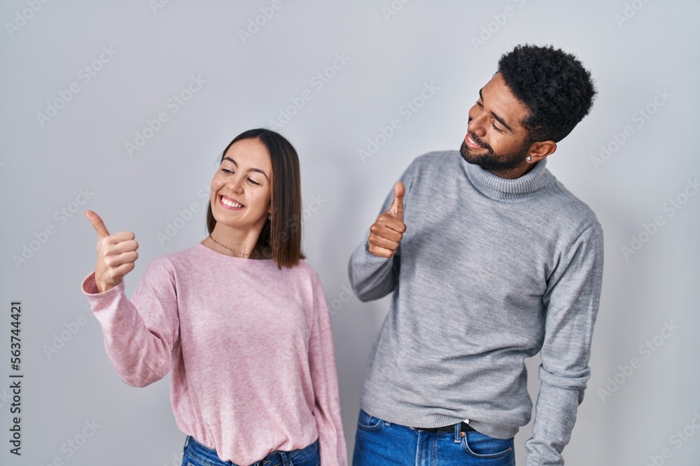 Young hispanic couple standing together looking proud, smiling doing thumbs up gesture to the side Young hispanic couple standing together looking proud, smiling doing thumbs up gesture to the side
