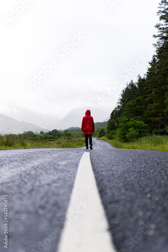 Portrait of a person wearing a red raincoat while walking alone on a road that crosses a valley surrounded by mountains and fir trees during a foggy day in the highlands, Scotland