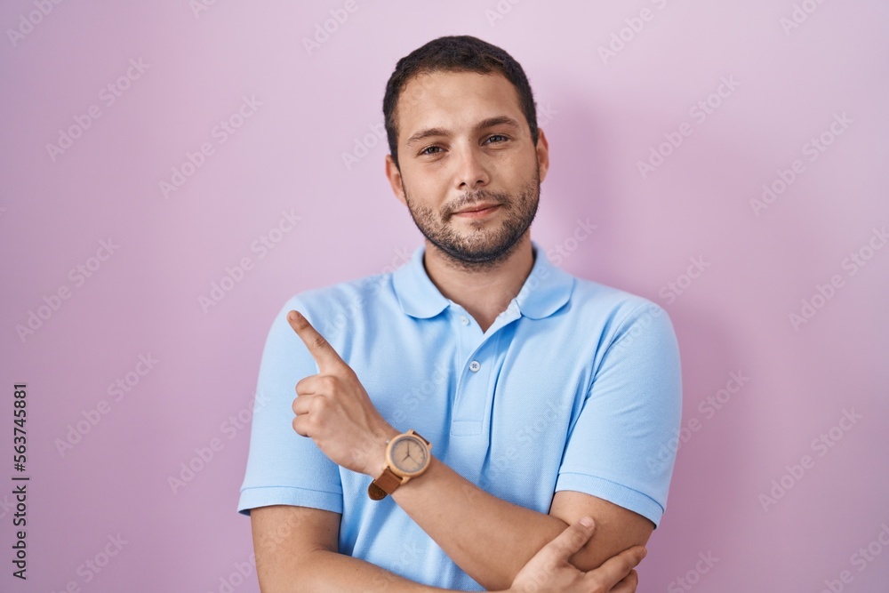 Hispanic man standing over pink background pointing with hand finger to the side showing advertisement, serious and calm face Hispanic man standing over pink background pointing with hand finger to the side showing advertisement, serious and calm face