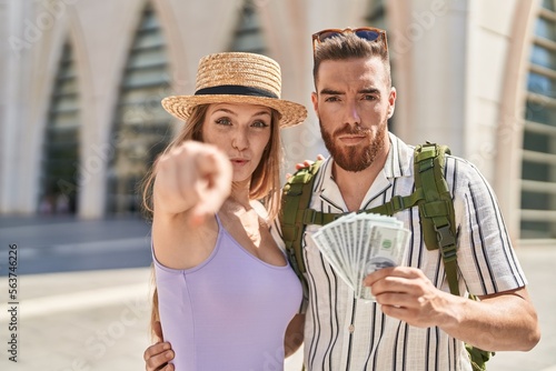 Young tourist couple holding dollars banknotes pointing with finger to the camera and to you, confident gesture looking serious