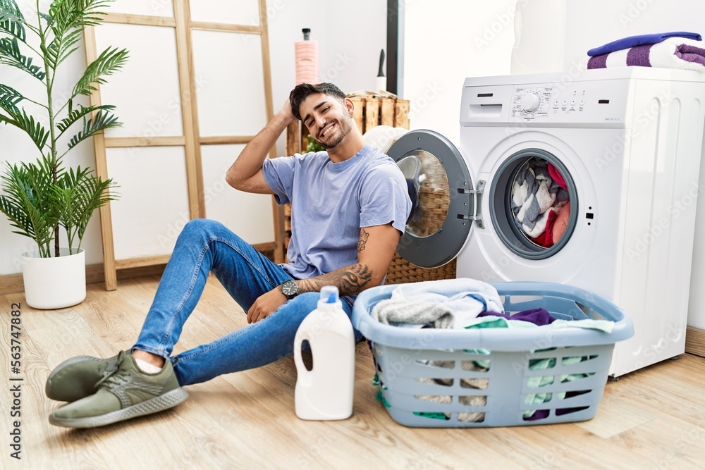 Young hispanic man putting dirty laundry into washing machine smiling confident touching hair with hand up gesture, posing attractive and fashionable Young hispanic man putting dirty laundry into washing machine smiling confident touching hair with hand up gesture, posing attractive and fashionable