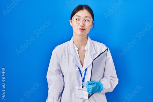 Chinese young woman working at scientist laboratory smiling looking to the side and staring away thinking.