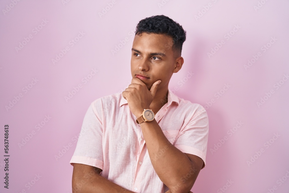 Young hispanic man standing over pink background with hand on chin thinking about question, pensive expression. smiling and thoughtful face. doubt concept. Young hispanic man standing over pink background with hand on chin thinking about question, pensive expression. smiling and thoughtful face. doubt concept.