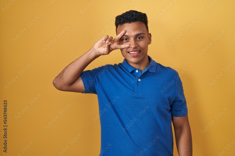 Young hispanic man standing over yellow background doing peace symbol with fingers over face, smiling cheerful showing victory Young hispanic man standing over yellow background doing peace symbol with fingers over face, smiling cheerful showing victory