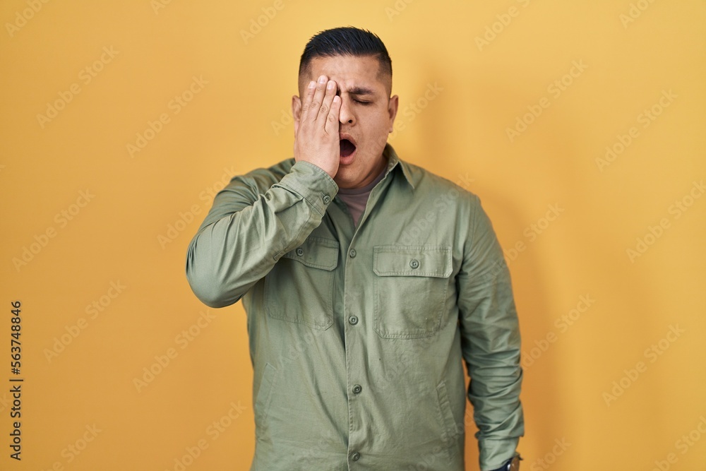 Hispanic young man standing over yellow background yawning tired covering half face, eye and mouth with hand. face hurts in pain. Hispanic young man standing over yellow background yawning tired covering half face, eye and mouth with hand. face hurts in pain.