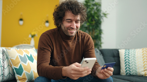 Young hispanic man using smartphone and credit card sitting on sofa at home